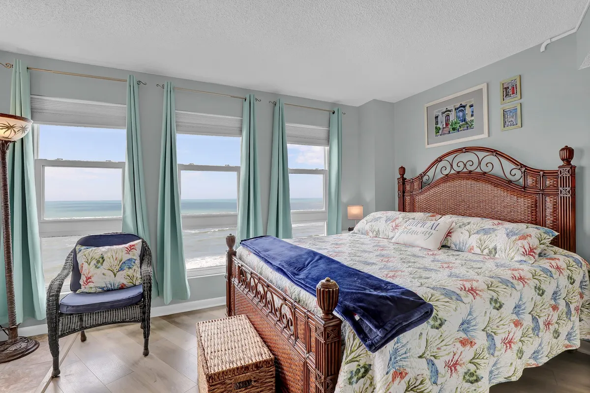 A serene master bedroom with a king bed adorned in floral bedding, large windows that frame an ocean view, a woven chair, and a woven storage basket. The room is filled with soft natural light, offering an inviting sanctuary for rest and privacy.