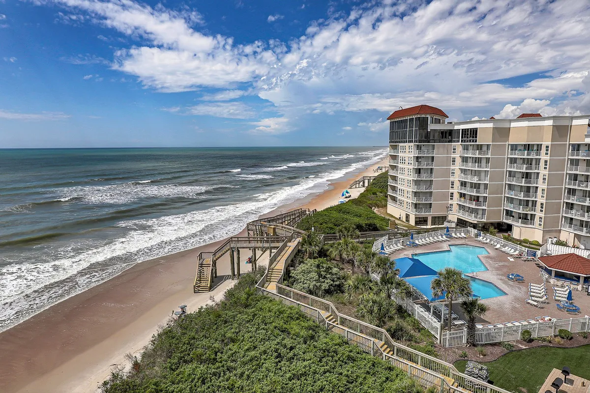 The hero image showcases the condo's stunning oceanfront location with sweeping views of the Atlantic, a brand new covered saltwater pool in the foreground, a sun-dappled pathway leading directly to the sandy beach, and inviting spaces for sunbathing and outdoor dining. The residential building stands elegantly, promising comfort just steps from the water's edge.