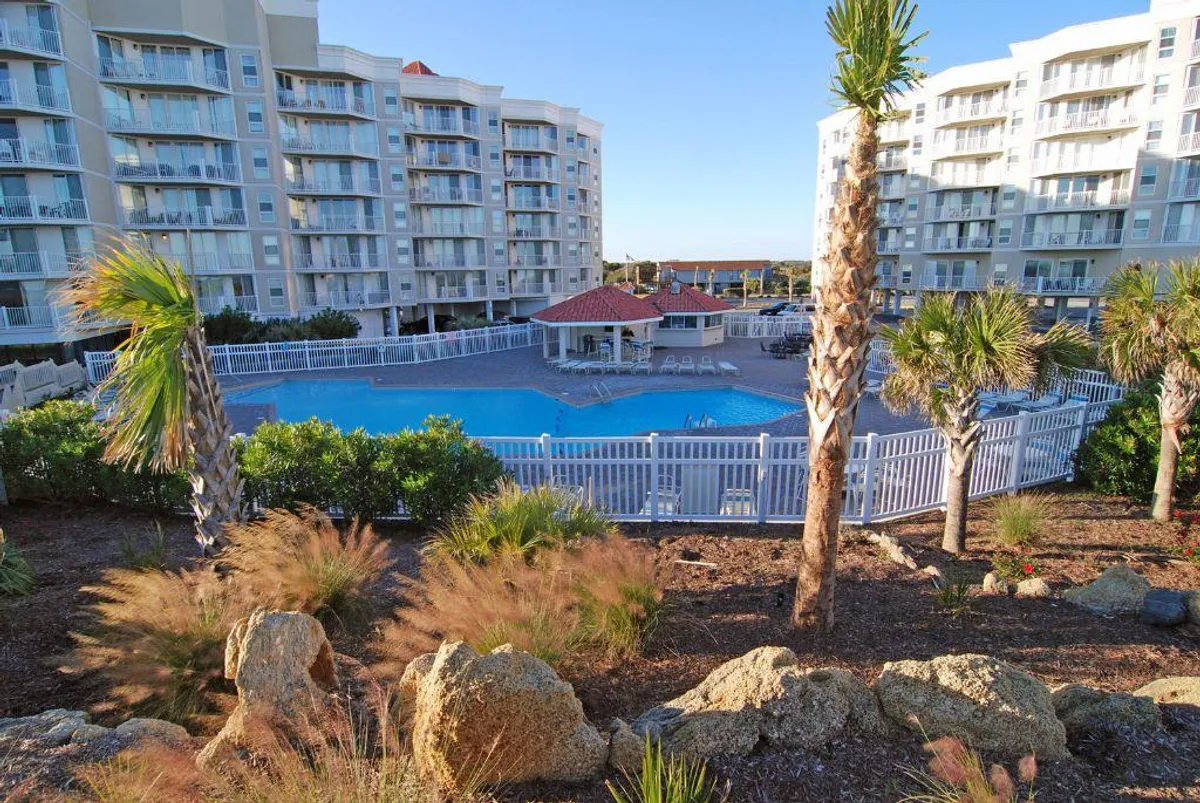 A sparkling swimming pool framed by swaying palm trees and resort buildings, with a lounge area featuring chairs for relaxation. The atmosphere is leisurely and welcoming, inviting guests to swim, sunbathe, or socialize poolside.