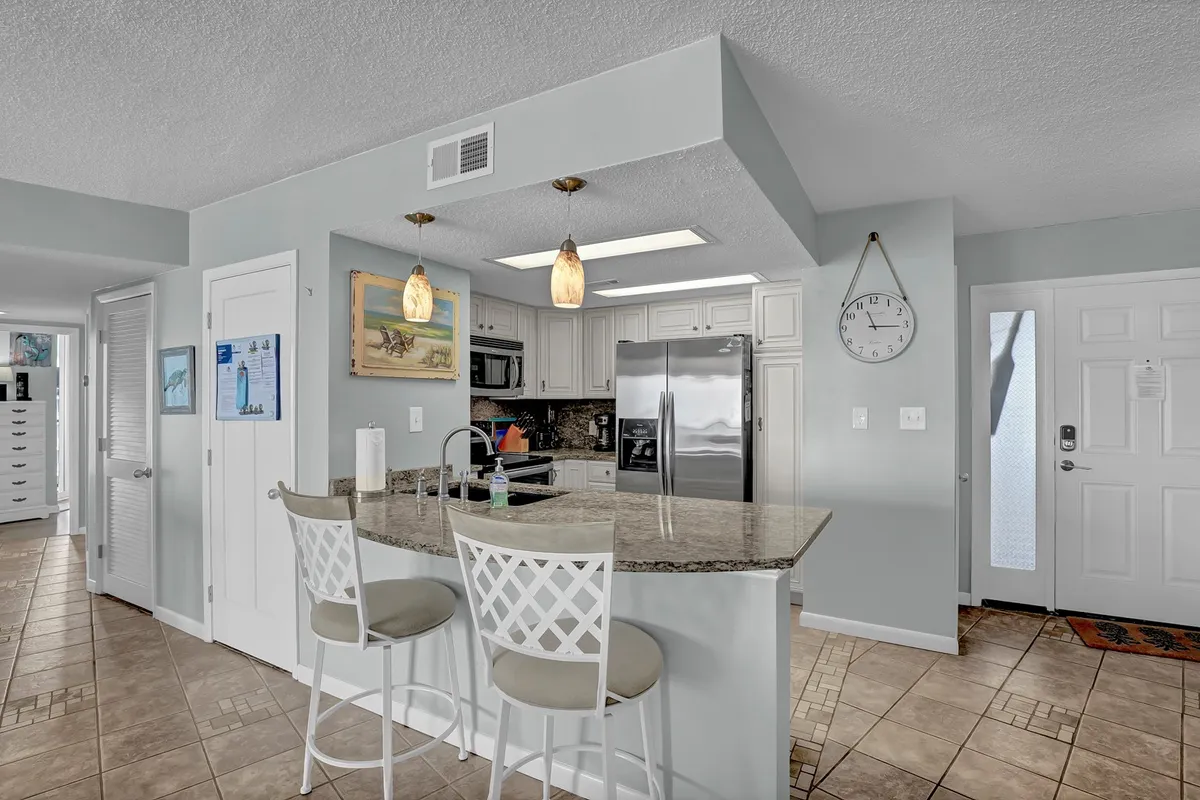 A modern kitchen space with a granite countertop, bar stools, refrigerator, and wall art. The setup is functional and stylish, perfect for cooking, socializing, or unwinding with a morning coffee.