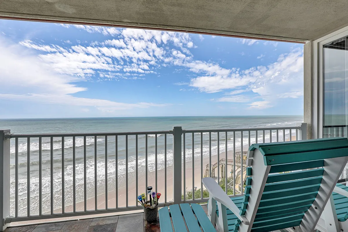 This exterior image features a serene ocean view captured from a private balcony, with the beach and cloudy sky stretching out in the distance. The balcony’s railing is visible, suggesting a perfect spot to relax while enjoying the coastal scenery. The mood is tranquil, inviting guests to unwind while watching dolphins or taking in a sunrise or sunset.
