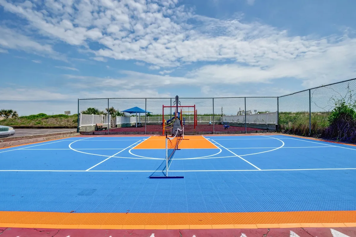 An energetic exterior scene showing a basketball court with a tennis net and a nearby playground structure. The area is designed for active group fun, with space for basketball games, tennis matches, and children’s play, surrounded by open sky.