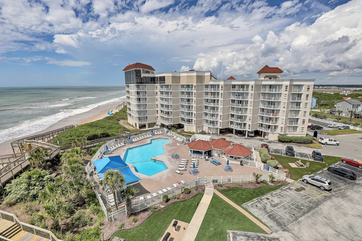A wide exterior shot showcasing the resort’s swimming pool, ocean view, and building architecture. Landscaped gardens frame the pool, while the beach and water are visible just beyond. The setting is vacation-like and serene, ideal for both swimming and social gatherings.