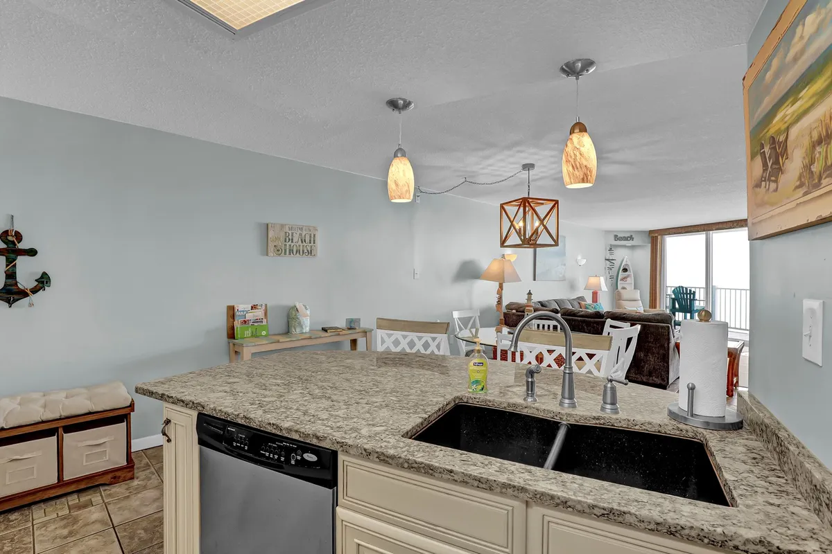 An inviting open-plan kitchen with a granite countertop, stylish pendant lights, and a cozy sitting area decorated with beach-themed accents. The space is well-lit and designed for both meal preparation and casual socializing, reflecting a fully equipped kitchen ready for extended stays.