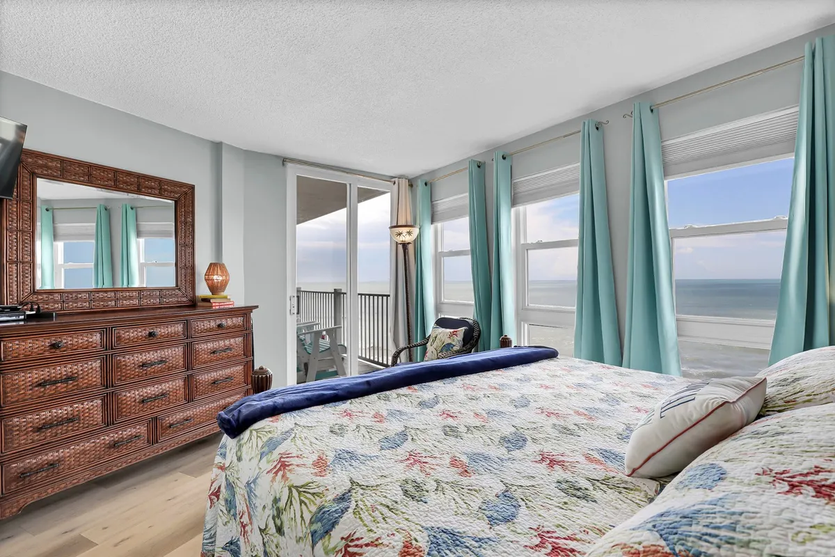 A serene bedroom featuring a king-sized bed with a floral quilt, a large dresser with a mirror, and windows offering a view of the ocean. The room is bright and inviting, with a focus on comfort and the option for personalized linen arrangements.