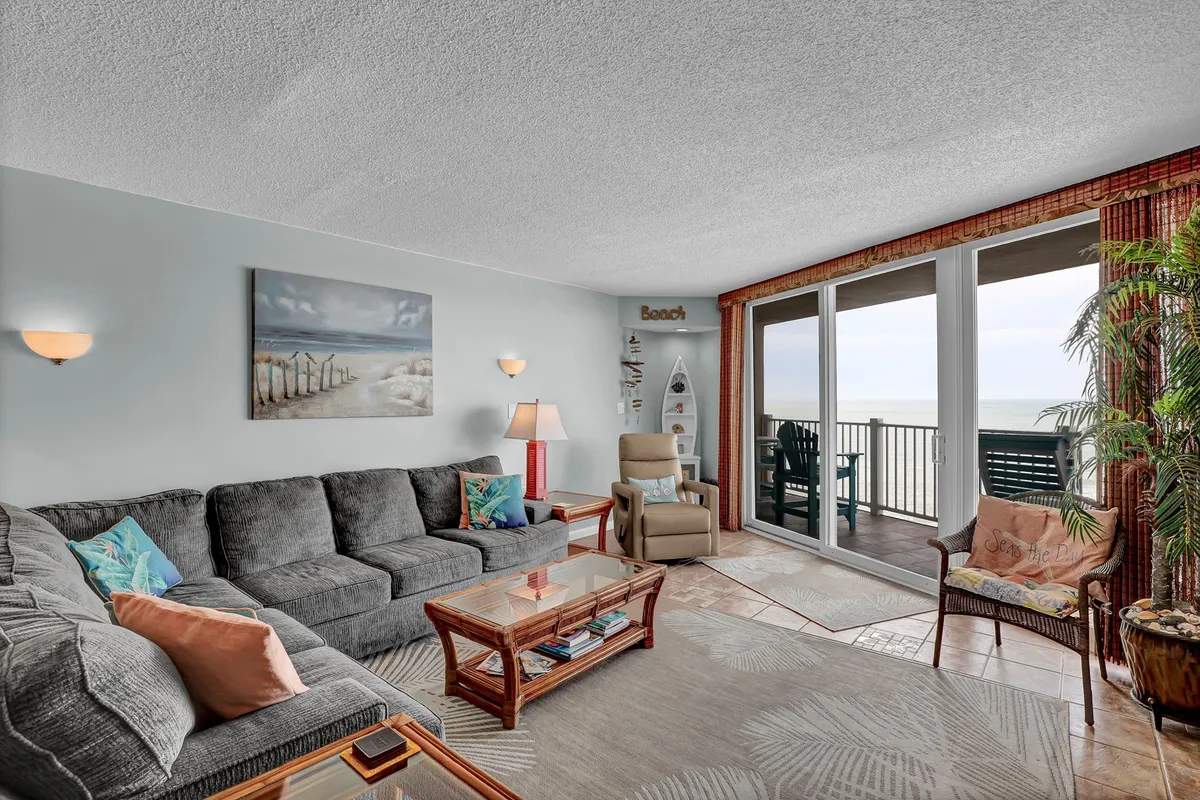 A comfortable living room with a large sectional sofa faces glass doors that offer sweeping ocean views and access to a private deck. The space is decorated with coastal artwork and features a coffee table, inviting guests to relax, socialize, or head outside to enjoy the salty breeze.