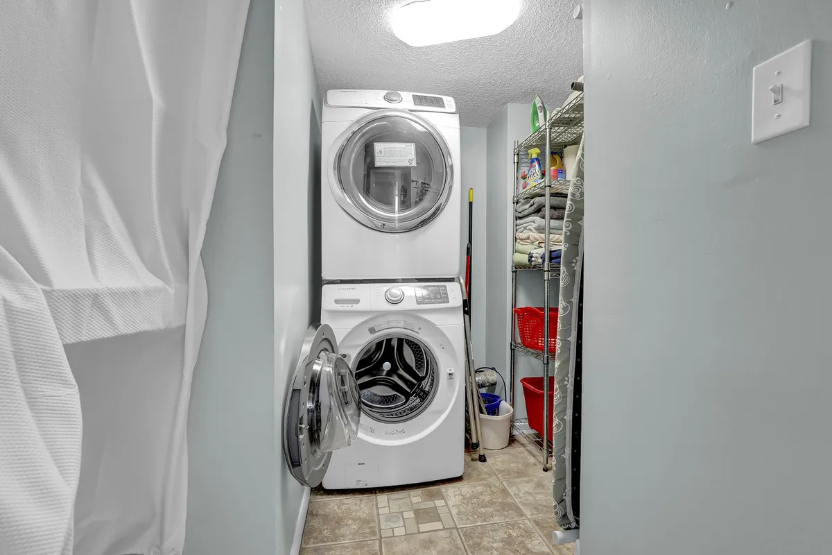 A practical laundry nook featuring a stacked washer and dryer, shelving unit, and laundry supplies. The space is organized and efficient, designed to keep family routines smooth during vacation.