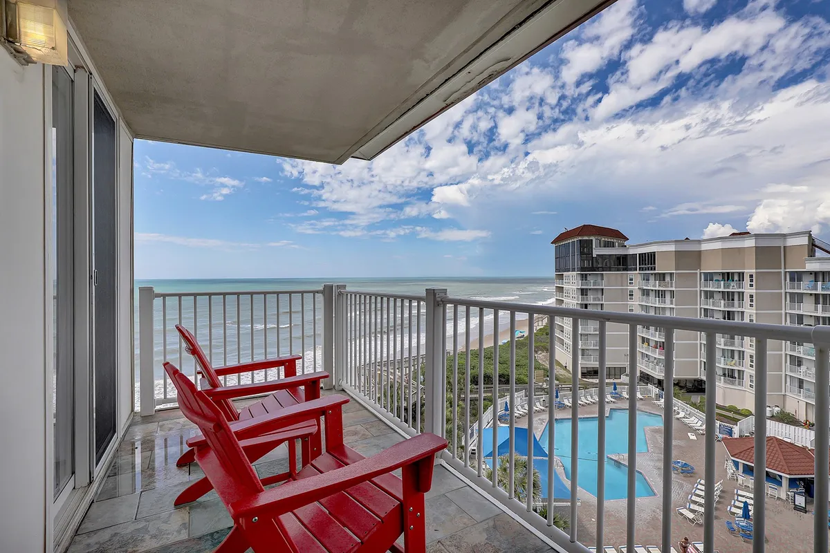 An exterior shot of a serene swimming pool area with red chairs overlooking both the sparkling pool and the expansive ocean beyond. The setting feels secluded and ideal for relaxation or small social gatherings.